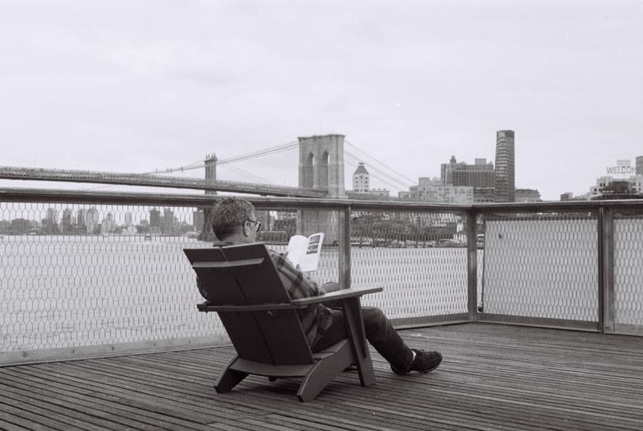 Black-and-white photo of a man reading in a deck chair facing the Brooklyn Bridge