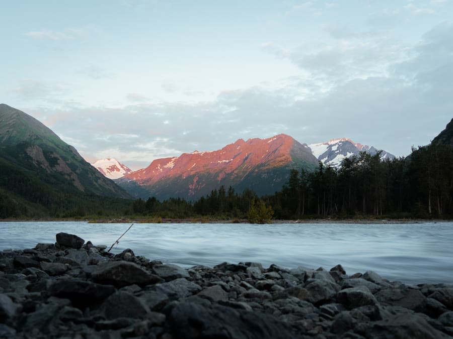 Alpenglow on a mountain range above a glacial river and rocky shore