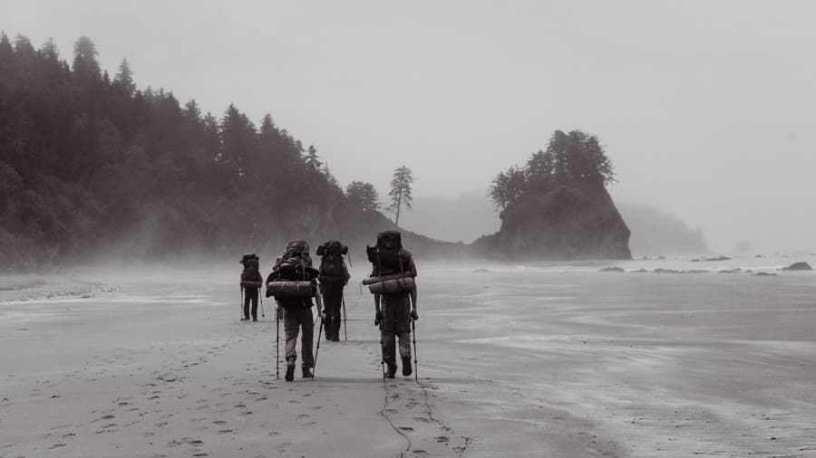 Four hikers with packs crossing a misty beach toward a sea stack